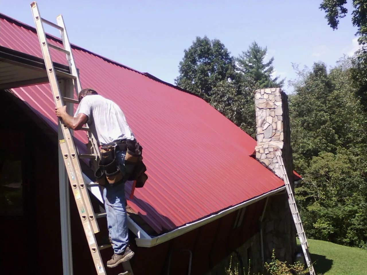 Roofer on a ladder beside a red metal roof and stone chimney — Soffit & Fascia Repair by Roofing Brothers in Valley Stream
