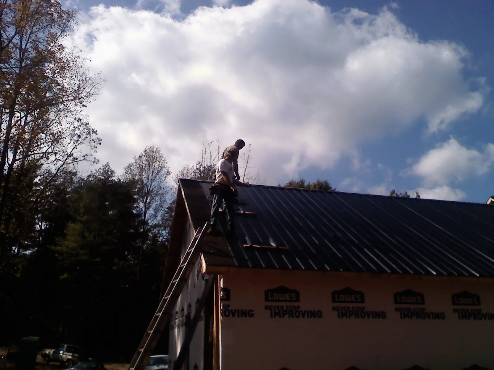 Roofer fastening dark metal panels high against a cloudy sky, from a Roof Inspection job in Valley Stream